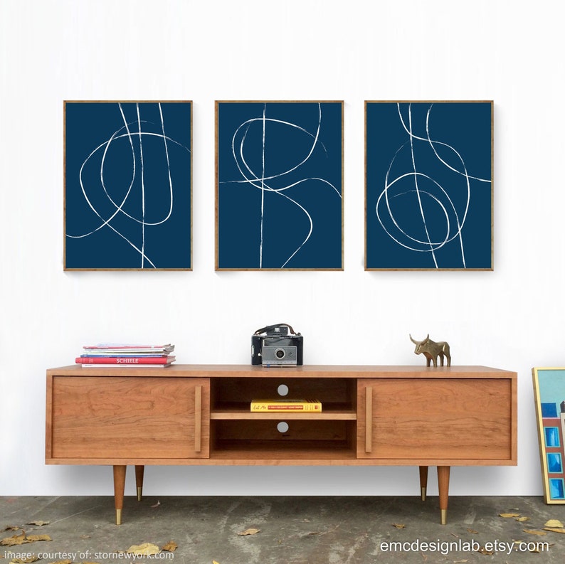 May include: Three framed abstract art prints with white lines on a dark blue background. The prints are hanging on a white wall above a wooden media console.
