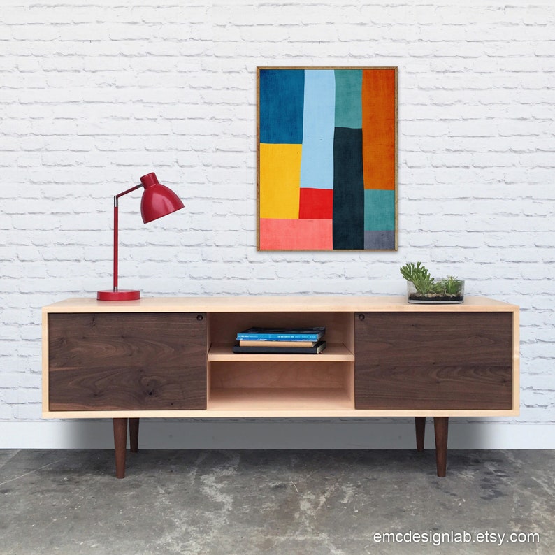 May include: A mid-century modern walnut and maple wood media console with sliding doors and a shelf. The console has a red lamp and a framed abstract art print with yellow, blue, red, and orange colors on a white brick wall.