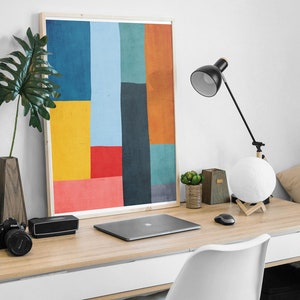 May include: A framed abstract art print with geometric shapes in shades of blue, yellow, red, orange, and green. The print is hanging on a white wall above a wooden desk with a laptop, a lamp, and a white chair.