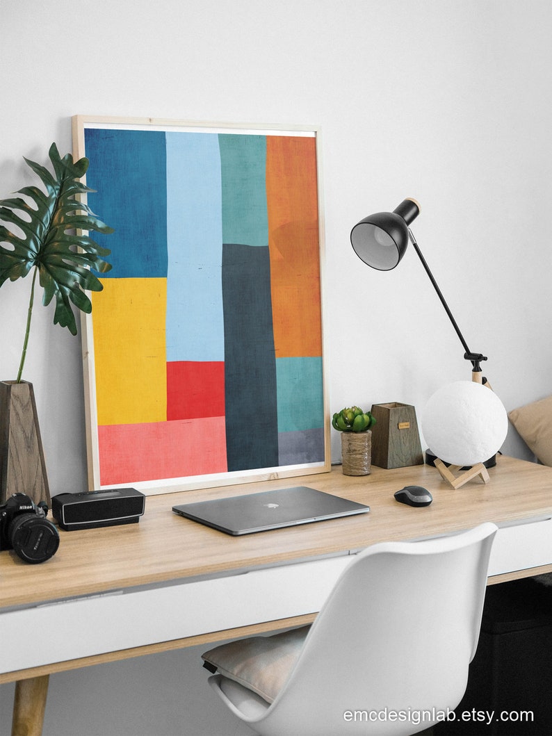 May include: A framed abstract art print with geometric shapes in shades of blue, yellow, red, orange, and green. The print is hanging on a white wall above a wooden desk with a laptop, a lamp, and a white chair.