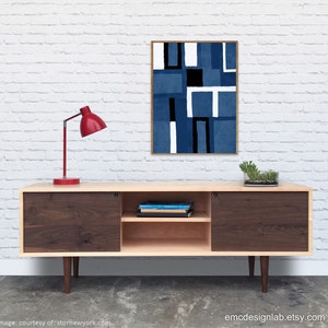 May include: A mid-century modern walnut and maple media console with sliding doors and a shelf. The console has a red lamp on top and a framed abstract art print with blue, white, and black geometric shapes hanging on the wall behind it.