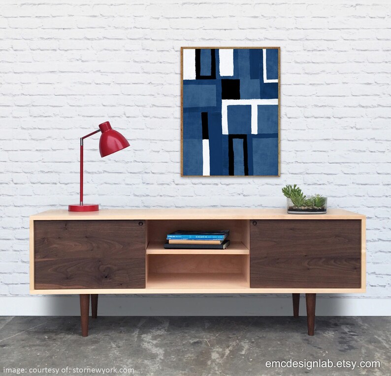 May include: A mid-century modern walnut and maple media console with sliding doors and a shelf. The console has a red lamp on top and a framed abstract art print with blue, white, and black geometric shapes hanging on the wall behind it.