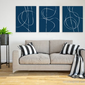 May include: Three abstract art prints with white lines on a dark blue background. The prints are hung on a white wall above a light gray couch with black and white striped pillows and a black and white throw blanket.