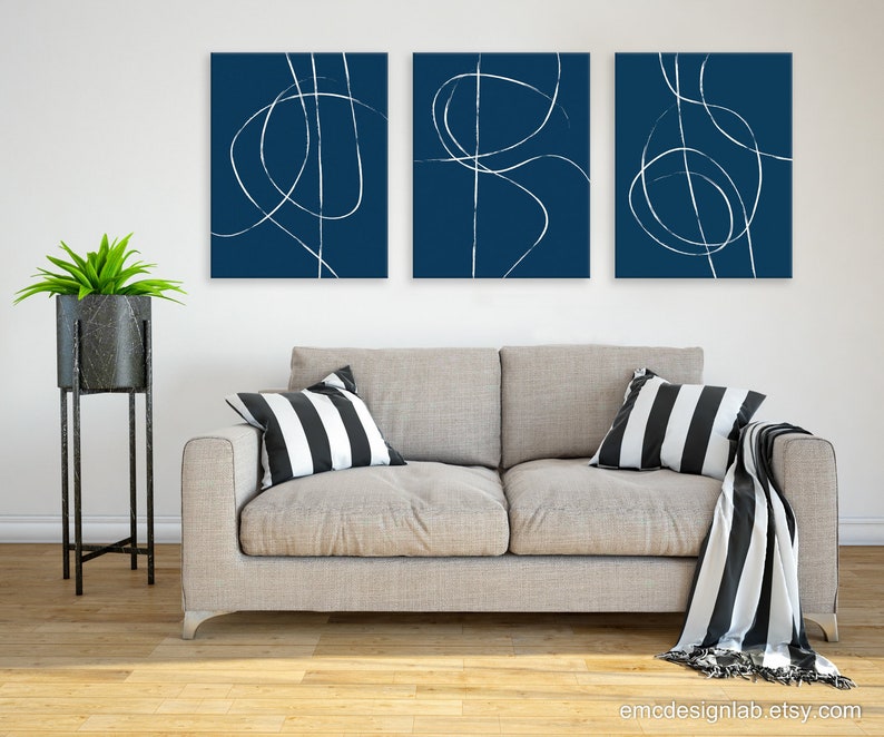 May include: Three abstract art prints with white lines on a dark blue background. The prints are hung on a white wall above a light gray couch with black and white striped pillows and a black and white throw blanket.