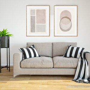 Pode incluir: Duas impress&otilde;es de arte abstrata emolduradas com formas geom&eacute;tricas e linhas em tons de rosa, cinza e branco, penduradas acima de um sof&aacute; cinza claro com almofadas e uma manta listrada em preto e branco.