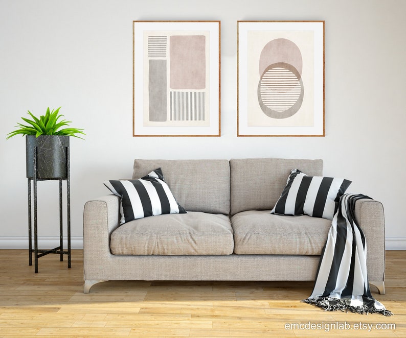 Pode incluir: Duas impress&otilde;es de arte abstrata emolduradas com formas geom&eacute;tricas e linhas em tons de rosa, cinza e branco, penduradas acima de um sof&aacute; cinza claro com almofadas e uma manta listrada em preto e branco.