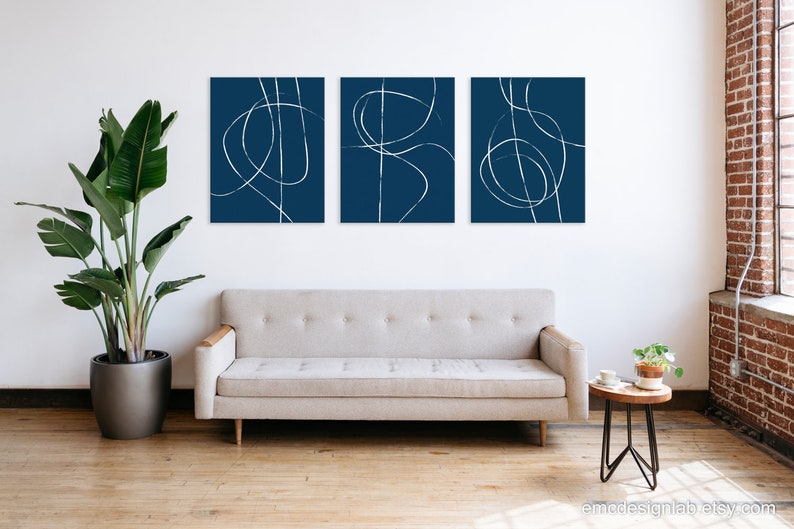 May include: Three abstract art prints with white lines on a dark blue background. The prints are arranged in a row on a white wall above a light gray couch.