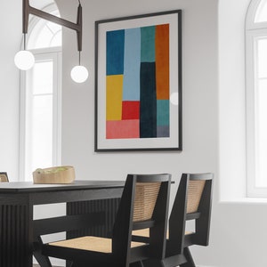May include: A black dining table with two black chairs with woven cane seats. The table has a wood grain pattern. A framed abstract art print with geometric shapes in yellow, red, blue, green, and orange hangs on the wall behind the table.