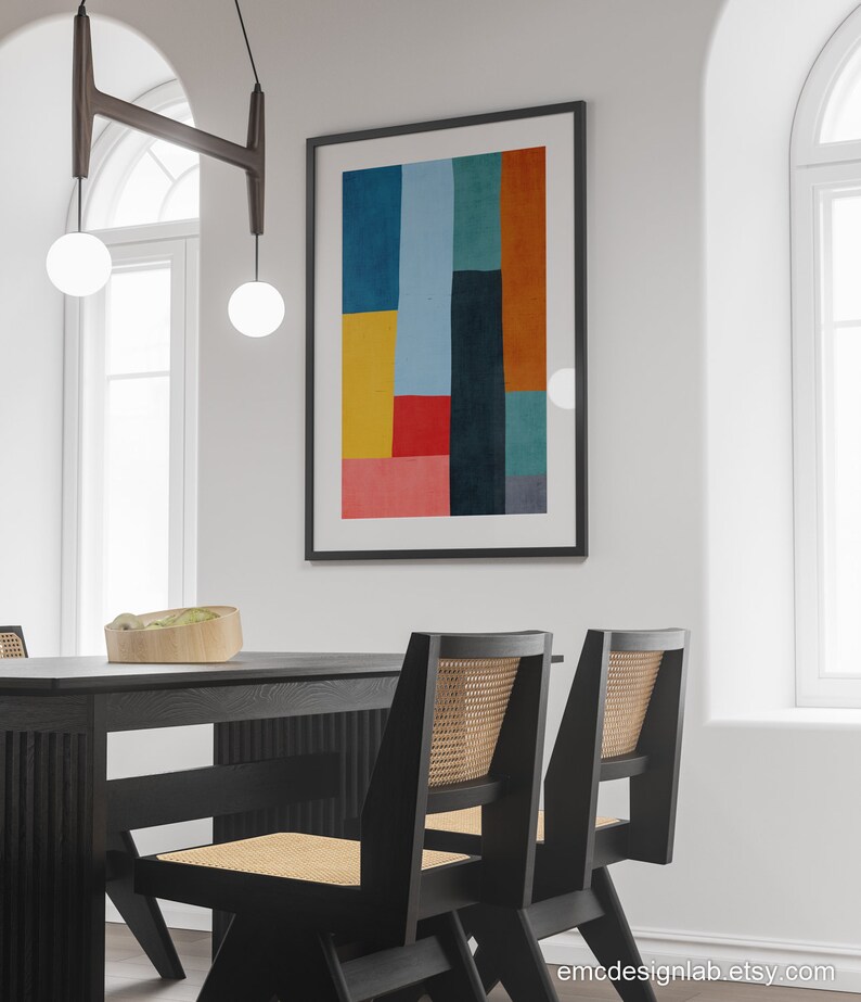 May include: A black dining table with two black chairs with woven cane seats. The table has a wood grain pattern. A framed abstract art print with geometric shapes in yellow, red, blue, green, and orange hangs on the wall behind the table.