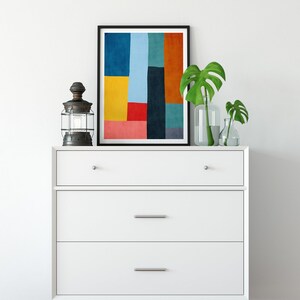 May include: A white dresser with three drawers and a black framed print of an abstract geometric design in shades of blue, yellow, red, orange, and green. The dresser has silver drawer pulls and sits on a wooden floor.