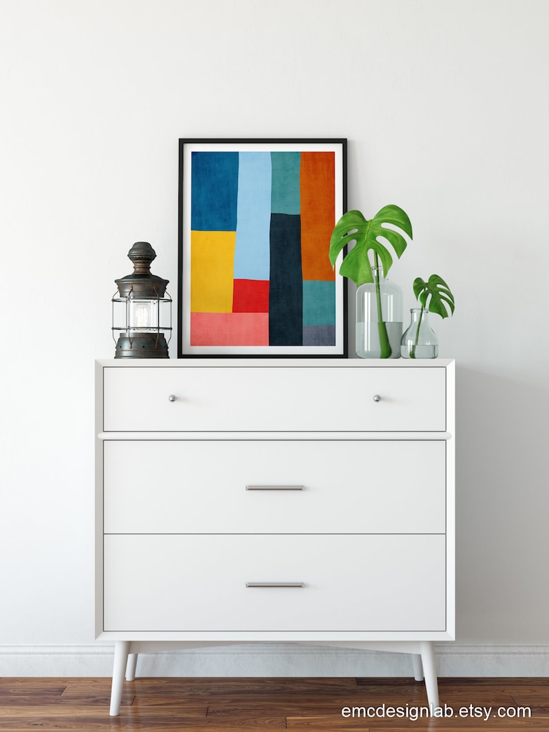 May include: A white dresser with three drawers and a black framed print of an abstract geometric design in shades of blue, yellow, red, orange, and green. The dresser has silver drawer pulls and sits on a wooden floor.