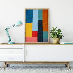 May include: A white mid-century modern sideboard with three drawers and two sliding doors. A teal blue desk lamp with a white shade is on the left side of the sideboard. A framed abstract print with geometric shapes in red, yellow, blue, green, and orange is centered above the sideboard. A potted plant is on each side of the sideboard.
