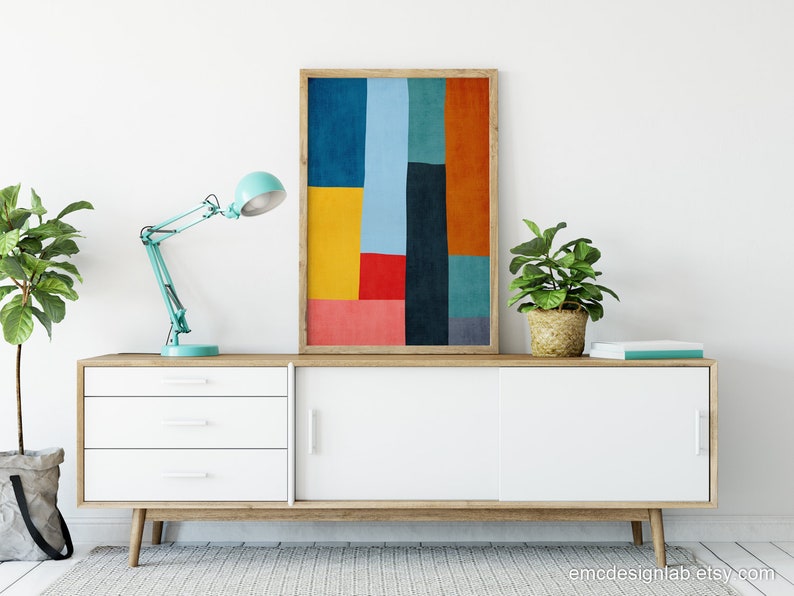 May include: A white mid-century modern sideboard with three drawers and two sliding doors. A teal blue desk lamp with a white shade is on the left side of the sideboard. A framed abstract print with geometric shapes in red, yellow, blue, green, and orange is centered above the sideboard. A potted plant is on each side of the sideboard.