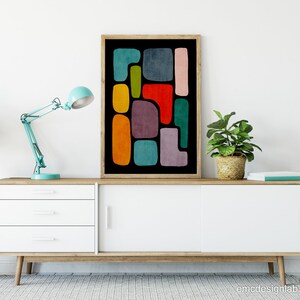 May include: A framed abstract art print with colorful geometric shapes on a black background. The print is displayed on a white dresser with a blue lamp and two potted plants.