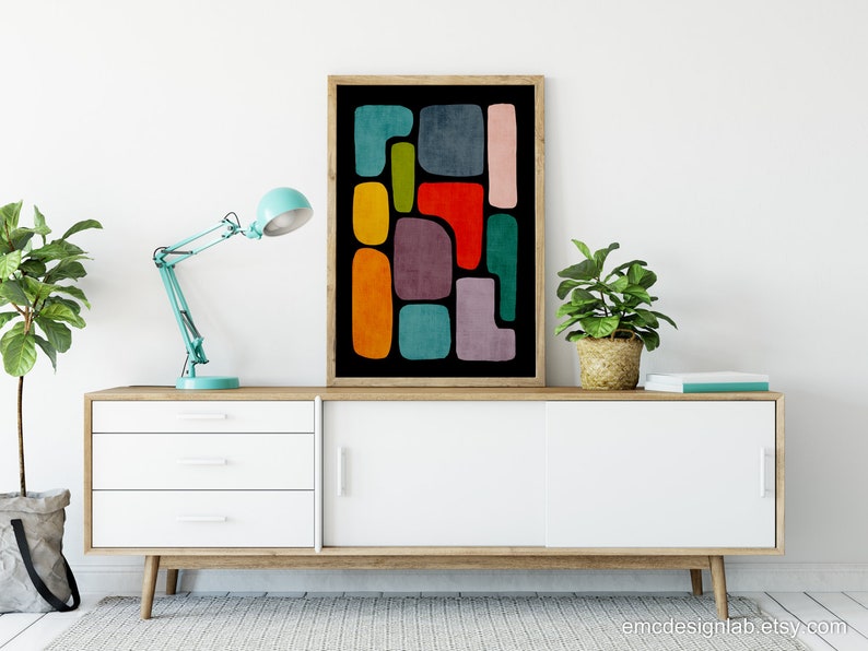 May include: A framed abstract art print with colorful geometric shapes on a black background. The print is displayed on a white dresser with a blue lamp and two potted plants.