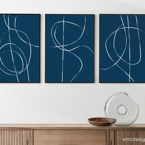 May include: Three abstract art prints with white lines on a dark blue background. The prints are framed in black and hung on a white wall above a wooden cabinet.
