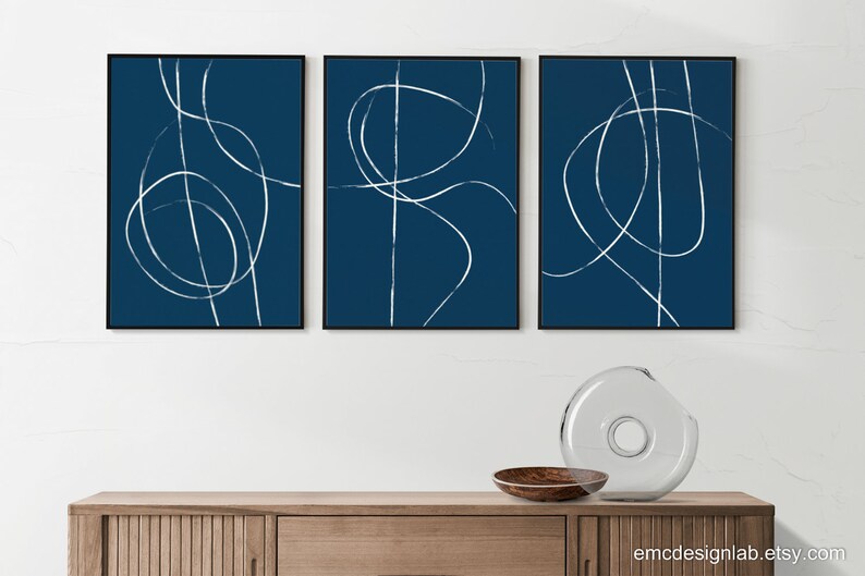May include: Three abstract art prints with white lines on a dark blue background. The prints are framed in black and hung on a white wall above a wooden cabinet.