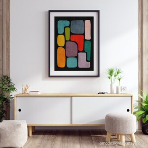 May include: Abstract art print with colorful geometric shapes on a black background. The print is framed in black and hanging on a white wall above a wooden cabinet.