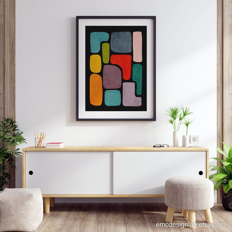 May include: Abstract art print with colorful geometric shapes on a black background. The print is framed in black and hanging on a white wall above a wooden cabinet.