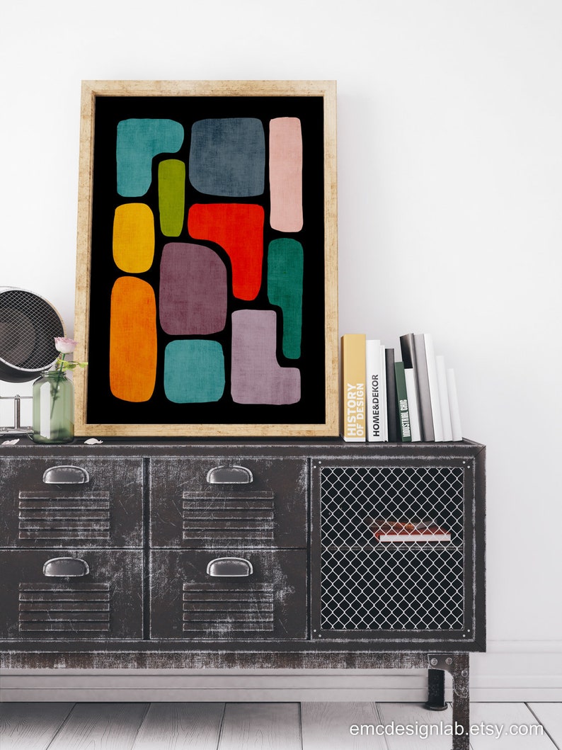 May include: Abstract geometric art print with colorful shapes on a black background. The print is framed in a light wood frame and is displayed on a metal cabinet with drawers.