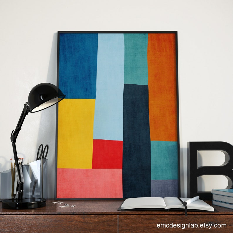 May include: Abstract art print with a geometric design featuring a variety of colors including yellow, red, blue, green, and orange. The print is framed in a black frame and is displayed on a desk with a lamp and other office supplies.