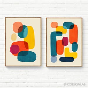 May include: Two framed abstract art prints. Each print features a beige background with overlapping shapes in orange, yellow, teal, and magenta. The prints are in wooden frames and are displayed against a white brick wall.