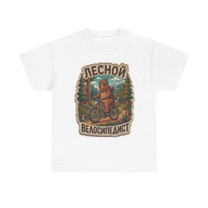 Peut inclure: Un t-shirt blanc avec une illustration centrale représentant un ours brun portant un bonnet et des lunettes de soleil, faisant du vélo dans une forêt. Le dessin inclut des pins, des montagnes et le texte cyrillique LЕSNOY VЕLOSIPЕDIST.