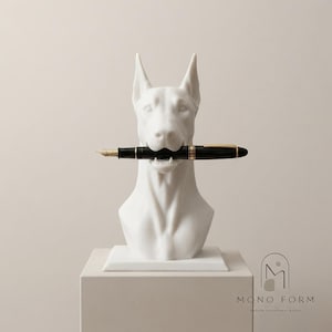 May include: A white Doberman dog bust sculpture featuring a black fountain pen with gold accents held horizontally in its mouth. The sculpture is mounted on a square base and includes the text MONO FORM at the bottom right corner.