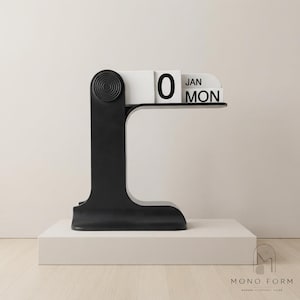 Peut inclure: Un calendrier de bureau noir en forme de L avec un détail latéral texturé circulaire et des blocs blancs affichant la date 0, le mois JAN et le jour MON. Le calendrier est posé sur une base rectangulaire blanche avec le texte MONO FORM.