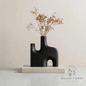 Puede incluir: Un jarrón de cerámica negro mate con una silueta escultural de múltiples arcos, que contiene un ramo de flores secas de color beige. La base del jarrón es rectangular y el texto MONO FORM MODERN SCULPTURAL DECOR es visible en la esquina.
