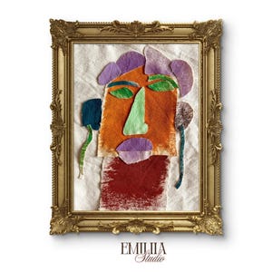 May include: A fabric collage portrait of a face in a gold ornate frame. The face features an orange head, green nose, purple hair and lips, and green eyes. A dark red neck section sits below. The text EMILIIA Studio is printed below the frame.