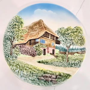 May include: A round ceramic decorative plate featuring a raised, three-dimensional relief scene of a rustic house with a thatched roof, stone foundation, and wooden walls, set in a landscape with green trees, a dirt path, and a blue sky.