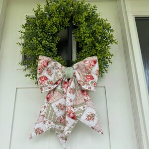 Könnte beinhalten: Ein runder, grüner Buchsbaumkranz mit einer großen Stoffschleife in der Mitte. Die Schleife besteht aus einem Stoff im Patchwork-Stil mit einem floralen Rosenmuster in Rot-, Rosa- und Grüntönen, akzentuiert durch einen kleinen hellgrünen Mittelknoten.