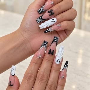 Könnte beinhalten: Lange, sargförmige Kunstnägel mit nudefarbener Basis, schwarzen French Tips, weißen Spinnennetzmustern, 3D-Geisterfiguren, kleinen schwarzen Schleifen und klaren Strasssteinen. Das Design besticht durch feine schwarze Linien.