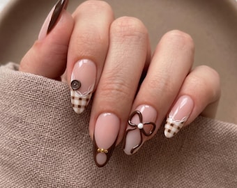 May include: Almond-shaped nails featuring a nude base with brown French tips, brown and white gingham patterns, a small brown button accent, a raised brown bow with a white center, and tiny gold beads on a dark brown accent nail.