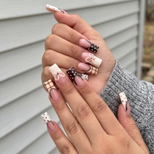 May include: Long square-shaped artificial nails featuring a mix of designs including brown and white polka dots, brown and white plaid patterns, and raised white bow accents on a nude base with dark brown French tips.