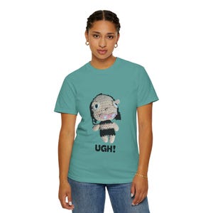 May include: A teal short-sleeve t-shirt featuring a graphic of a crocheted doll with dark hair, a beige face, and a black and beige striped body. The doll has blue eyes and a pink mouth. The text UGH! is printed in black below the doll graphic.