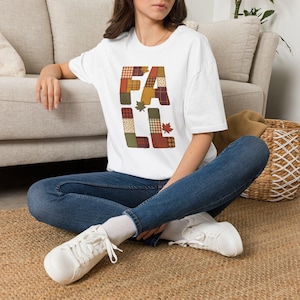 May include: A white short-sleeved t-shirt featuring the word FALL printed in large block letters. Each letter is filled with a patchwork pattern of plaid and solid fabrics in autumn colors like orange, brown, and green, accented by maple leaves.