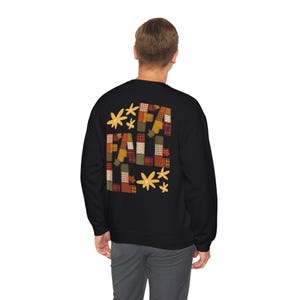 May include: A black long-sleeve sweatshirt featuring a graphic on the back with the word FALL in a patchwork plaid pattern of orange, red, green, and cream colours. The design includes stylised leaf illustrations in yellow and orange tones.