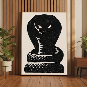 May include: A rectangular wall art print featuring a high-contrast black graphic of a coiled cobra with a flared hood. The snake has two sharp, light-coloured almond-shaped eyes set against a solid off-white background within a thin wooden frame.