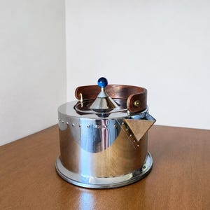 Peut inclure: Une bouilloire cylindrique en acier inoxydable avec une finition miroir polie, dotée d'une poignée en bois incurvée, d'un petit bec triangulaire et d'un couvercle central surmonté d'un petit bouton rond en verre bleu foncé.