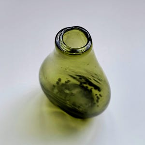 Può includere: Un piccolo vaso in vetro a forma di goccia di una tonalità verde oliva traslucida. Il recipiente presenta un'apertura circolare stretta nella parte superiore e un corpo liscio e arrotondato con sottili inclusioni vorticose verde scuro.