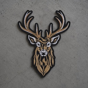 May include: An embroidered patch featuring the head of a deer with large antlers. The design uses shades of tan, brown, and white thread to create texture and detail, all outlined with a thick black border against a neutral gray background.
