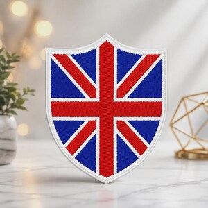 May include: A shield-shaped embroidered patch featuring the Union Jack flag design. The patch displays a red cross and diagonal stripes on a blue background, all bordered by a thick white stitched edge, creating a distinct heraldic appearance.