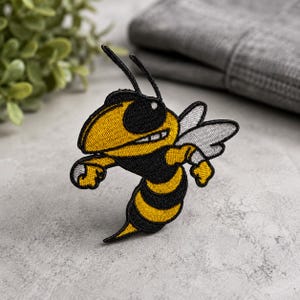 May include: An embroidered patch featuring a cartoon wasp with a yellow and black striped body, a large black eye, and white wings. The design includes a black outline and a determined facial expression with visible teeth, rendered in thread.