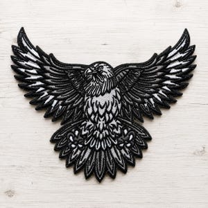 May include: An embroidered patch featuring a bald eagle in flight with wings fully spread. The design uses a monochromatic color palette of black, white, and shades of gray to define the feathers, head, and tail. The eagle is facing forward.