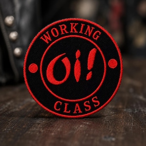 May include: A circular embroidered patch featuring a black background with a red border. The text "WORKING" is arched at the top and "CLASS" at the bottom in red. The center displays the red text "Oi!" with a red dot on the left and right sides.