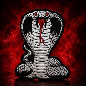 May include: An embroidered patch featuring a silver and black cobra with its hood flared. The snake has red eyes and a red forked tongue, with detailed scale patterns throughout its body. The design is outlined in a thick black border.