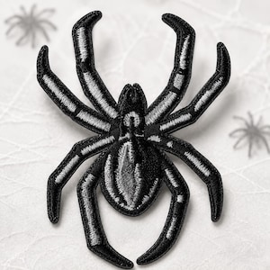 May include: An embroidered iron-on patch shaped like a spider, featuring a black thread base with silver-grey highlights along the eight legs and the central body. The design has a raised texture with a defined border and a satin stitch finish.
