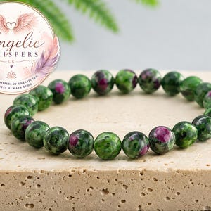 May include: A beaded bracelet featuring polished, spherical stones in shades of deep forest green and speckled magenta. The beads are strung together in a circular arrangement, showcasing natural colour variations and a smooth, reflective surface finish.